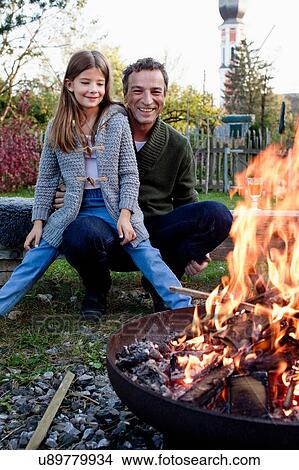 Picture - Girl sitting on father's lap in garden watching fire pit. Fotosearch