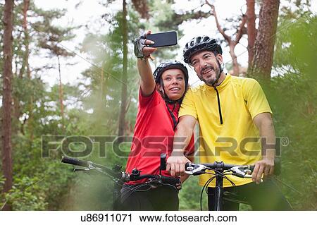 Stock Image - Happy mountain biking couple posing for smartphone selfie in forest. Fotosearch