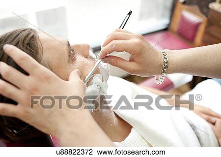 Stock Image - High angle over shoulder view of young man in barbershop being shaved with straight razor. Fotosearch