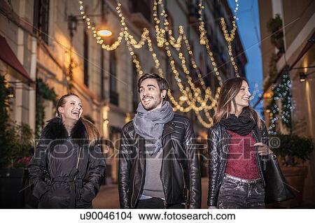 Picture - Low angle front view of friends walking down street in the evening smiling. Fotosearch
