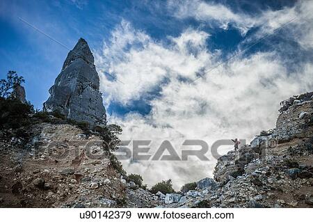 Low angle view of young man standing on cliff pointing at cone shape rock formation, Ogliastra, Sardinia, Italy View Large Photo Image Stock Photo - Low angle view of young man standing on cliff pointing at cone shape rock formation, Ogliastra, Sardinia, Italy. Fotosearch