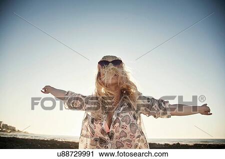 Stock Image - Low angle view of young woman arms open wearing sunglasses, hair covering face. Fotosearch