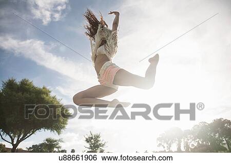 Stock Photograph - Low angle view of young woman jumping in mid air, arms raised. Fotosearch