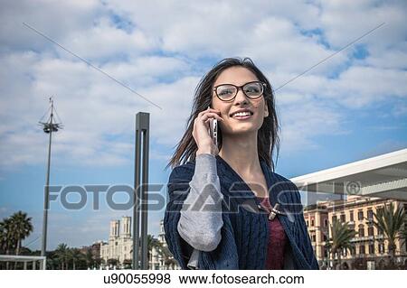 Stock Photo - Low angle view of young woman wearing eyeglasses using smartphone looking away smiling. Fotosearch