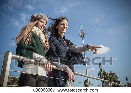 Stock Photo - Low angle view of young women leaning against railing looking away pointing and smiling. Fotosearch