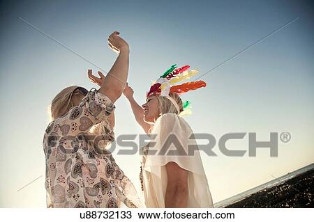 Stock Image - Low angle view of young women wearing feather headdress arms raised dancing in sunlight. Fotosearch