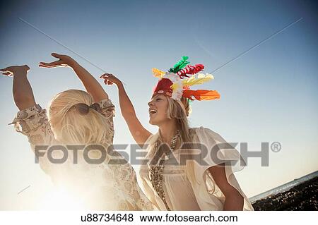 Stock Photo - Low angle view of young women wearing feather headdress and sunglasses with hair covering face dancing in sunlight. Fotosearch
