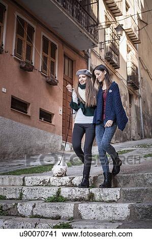 Stock Image - Low angle view of young women with dog walking down stone steps arms in arm smiling. Fotosearch