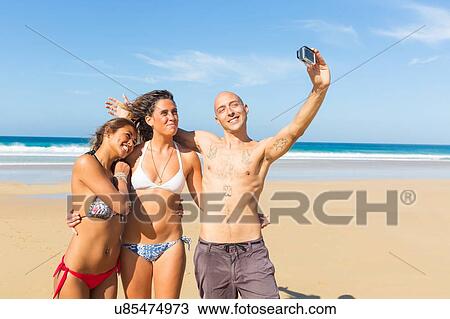 Man and two young women wearing swimwear taking selfie on beach, El Cotillo, Fuerteventura, Spain View Large Photo Image Stock Image - Man and two young women wearing swimwear taking selfie on beach, El Cotillo, Fuerteventura, Spain. Fotosearch
