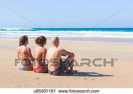 Stock Image - Man and two young women wearing swimwear looking to sea from beach, El Cotillo, Fuerteventura, Spain. Fotosearch