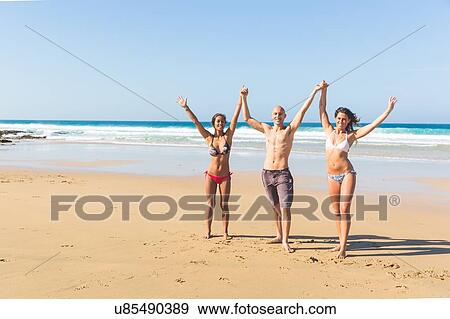 Stock Photo - Man and two young women wearing swimwear holding raised hands on beach, El Cotillo, Fuerteventura, Spain. Fotosearch
