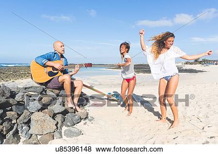 Stock Photo - Man playing acoustic guitar and women dancing on beach, El Cotillo, Fuerteventura, Spain. Fotosearch