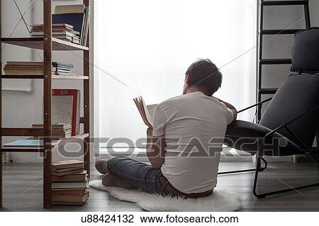 Stock Image - Man sitting on living room floor reading a book. Fotosearch