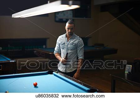 Stock Image - Man with beer standing at pool table. Fotosearch