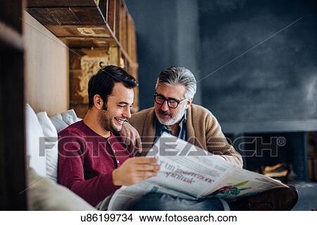 Mature man and adult son sitting on living room sofa reading newspaper View Large Photo Image Picture - Mature man and adult son sitting on living room sofa reading newspaper. Fotosearch