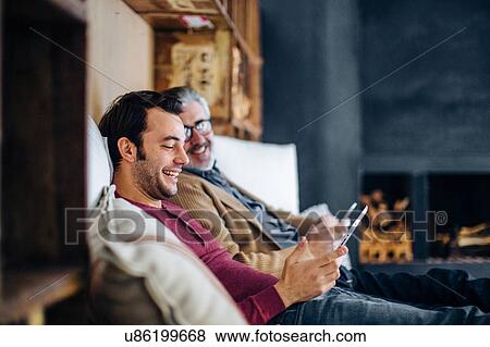 Stock Photo - Mature man and adult son sitting on living room sofa using digital tablet. Fotosearch