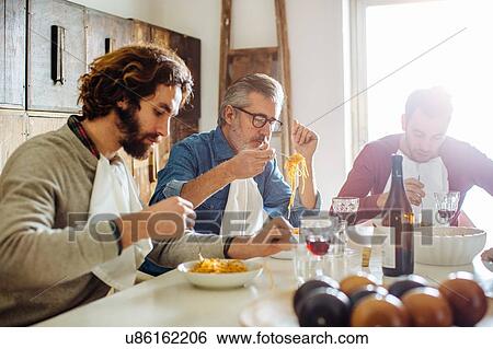 Stock Photograph - Mature man and adult sons eating spaghetti lunch at dining table. Fotosearch