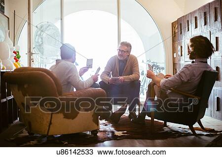 Stock Image - Mature man and adult sons sitting in living room looking at photographs. Fotosearch