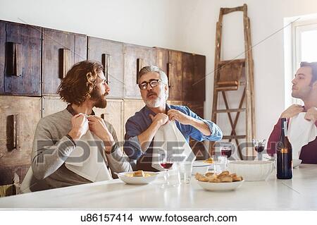 Picture - Mature man and adult sons tucking in napkins for lunch at dining table. Fotosearch