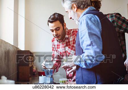 Stock Image - Mature man and two adult sons pouring coffee in kitchen. Fotosearch