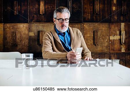 Stock Photography - Mature man reading smartphone text at dining table. Fotosearch