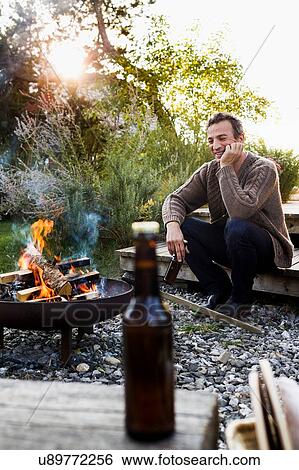 Stock Photograph - Mature man sitting by fire pit with beer, relaxing. Fotosearch
