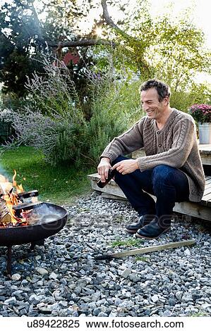 Stock Photography - Mature man sitting by fire pit with beer, relaxing. Fotosearch