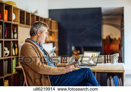 Mature man sitting in living room using smartphone and laptop View Large Photo Image Picture - Mature man sitting in living room using smartphone and laptop. Fotosearch