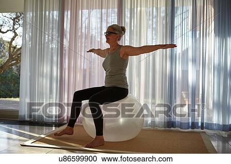 Stock Image - Mature woman balancing on exercise ball in front of patio doors. Fotosearch