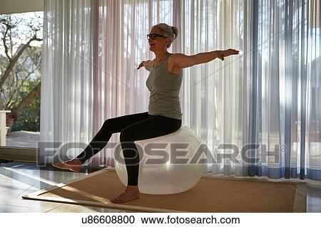 Stock Image - Mature woman balancing with leg raised on exercise ball in front of patio doors. Fotosearch