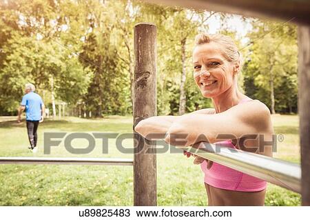 Stock Image - Mature woman in park leaning against metal bar looking at camera smiling. Fotosearch