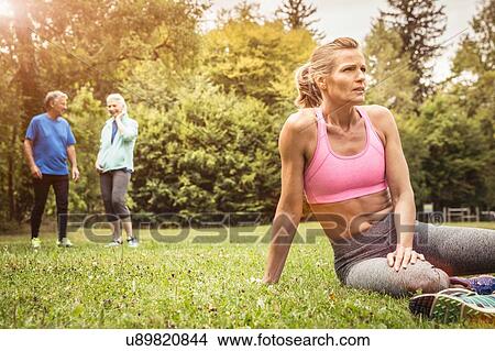 Picture - Mature women in park wearing sports clothing sitting on grass looking away. Fotosearch