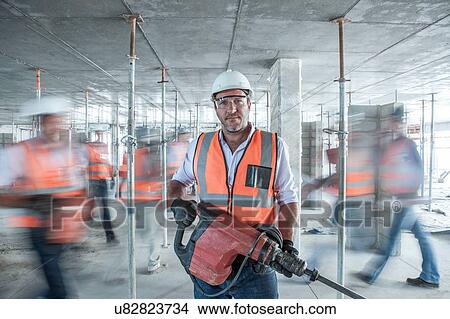 Mid adult male builder holding building equipment on busy construction site View Large Photo Image Picture - Mid adult male builder holding building equipment on busy construction site. Fotosearch