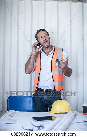 Mid adult male construction foreman talking on smartphone at desk in portable cabin View Large Photo Image Stock Photo - Mid adult male construction foreman talking on smartphone at desk in portable cabin. Fotosearch