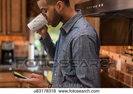 Stock Photo - Mid adult man in kitchen reading smartphone text and drinking coffee. Fotosearch