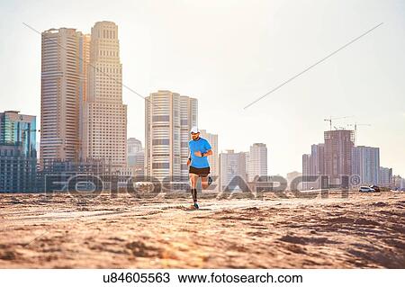 Stock Image - "Mid adult man running on sand by skyscrapers, Dubai, United Arab Emirates". Fotosearch
