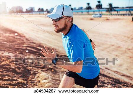 Stock Photo - "Mid adult man wearing baseball cap running up sand dune, Dubai, United Arab Emirates". Fotosearch