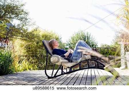 Stock Photography - Mid adult woman relaxing on lounge chair on wooden decking. Fotosearch