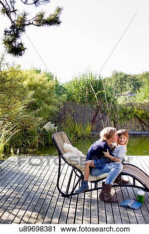 Stock Image - Mother and daughter hugging on lounge chair on decking. Fotosearch