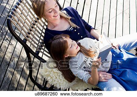 Stock Image - Mother and daughter relaxing on lounge chair, girl reading book. Fotosearch