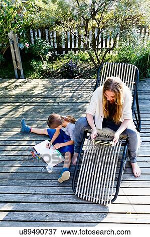 Stock Photo - Mother using tablet, daughter drawing on wooden decking. Fotosearch
