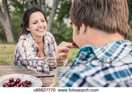 Stock Image - Over shoulder view of young woman feeding young man cherries at picnic table. Fotosearch