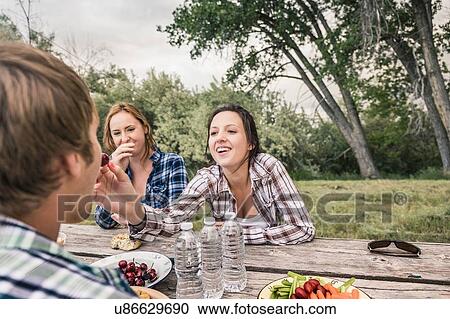 Stock Image - Over shoulder view of young women feeding young man cherries at picnic table. Fotosearch
