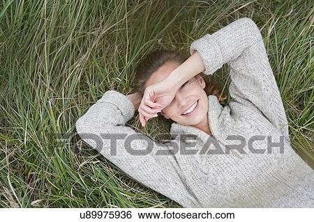 Stock Photograph - Overhead view of young woman lying in grass shielding eyes, looking at camera smiling. Fotosearch