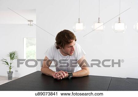 Stock Image - Pensive young man sitting at dining room table. Fotosearch