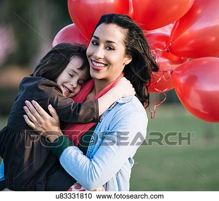 Stock Image - Portrait of girl with bunch of red balloons hugging mother in park. Fotosearch