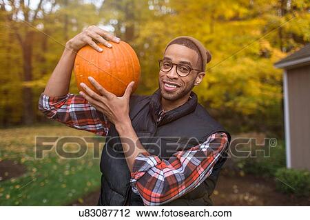 Stock Image - Portrait of mid adult man carrying pumpkin on shoulder in autumn garden. Fotosearch