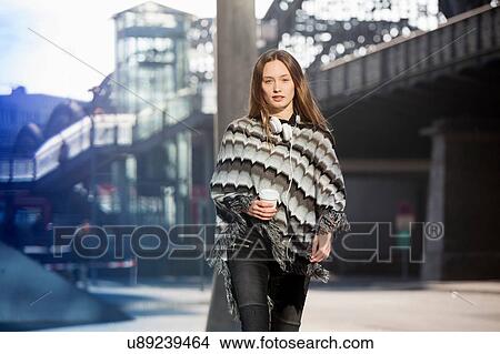Picture - Portrait of mid adult woman wearing poncho strolling in city with takeaway coffee. Fotosearch