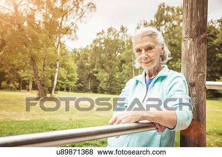 Stock Photo - Portrait of senior woman leaning against metal bar in park looking at camera smiling. Fotosearch