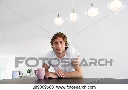 Stock Image - Portrait of serious young man sitting at dining room table. Fotosearch